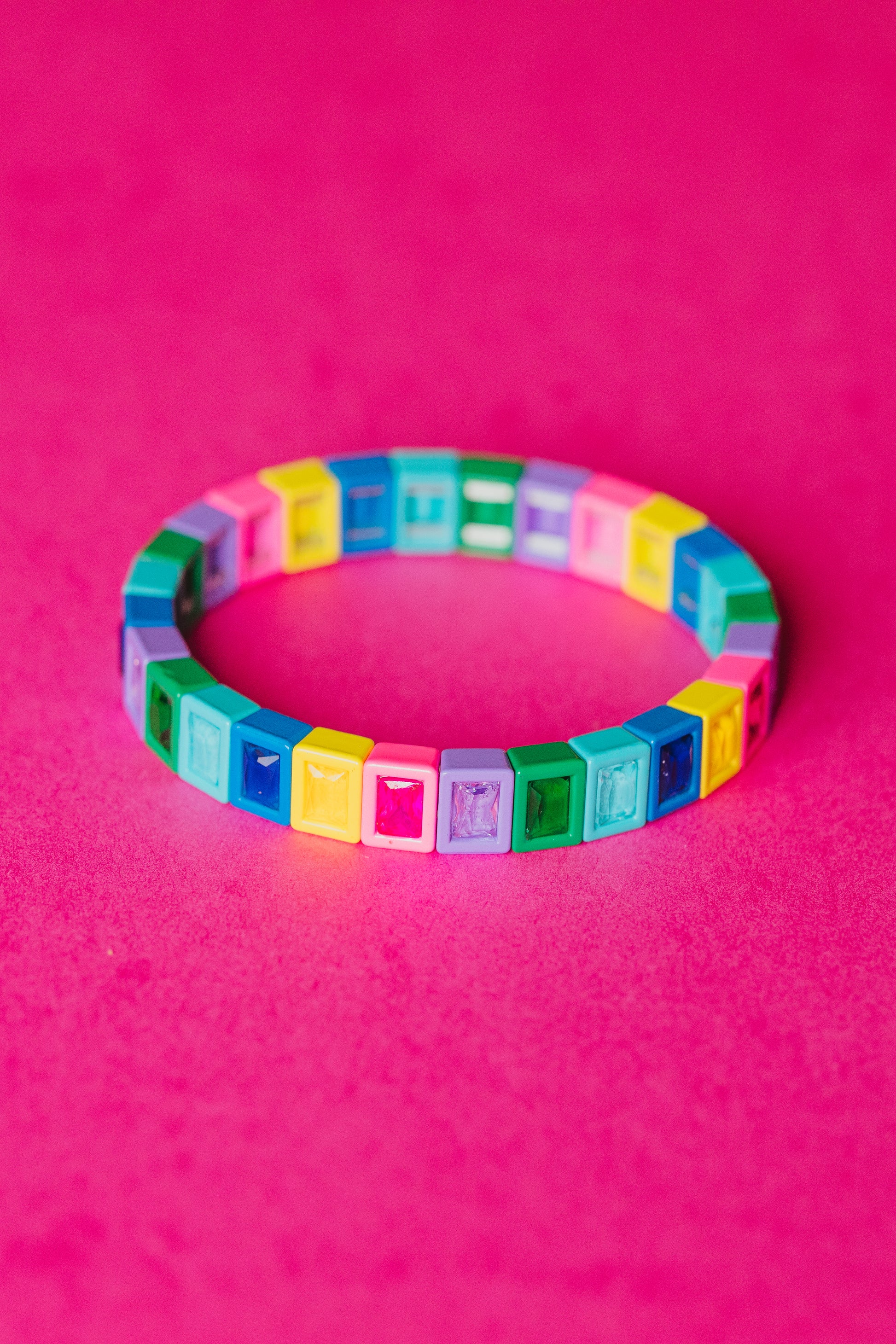 A colorful elastic bracelet made of small square plastic tiles arranged in a circle against a bright pink background. Each tile features a gem-like inset in various colors including yellow, blue, green, purple, pink, and turquoise. The bracelet is photographed from directly above.