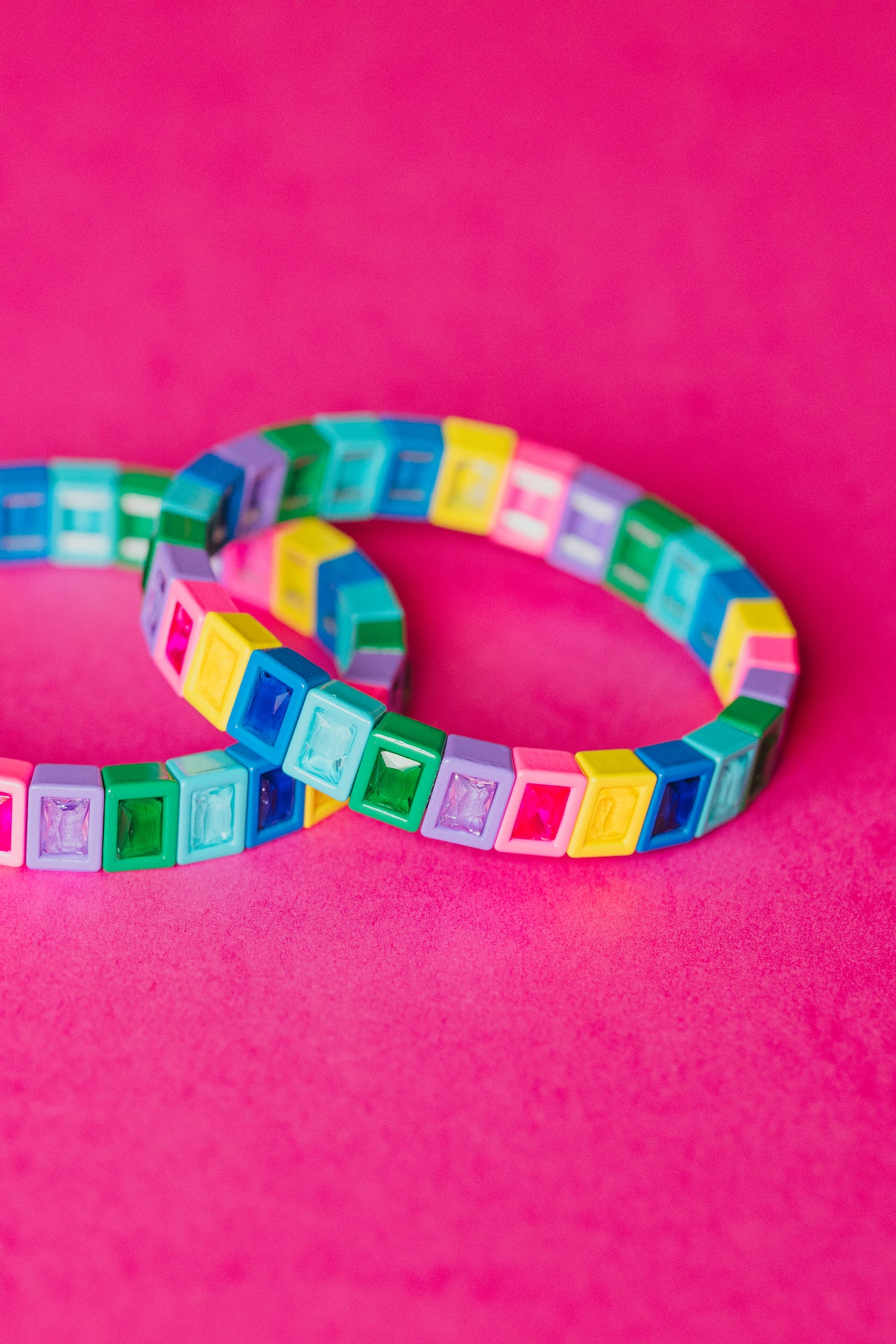 Two identical colorful tile bracelets overlapping on a bright pink background. Each bracelet is made of small square plastic tiles with gem-like insets in rainbow colors. The tiles are arranged to create a stretchy bracelet. The image shows the bracelet's construction and flexibility.