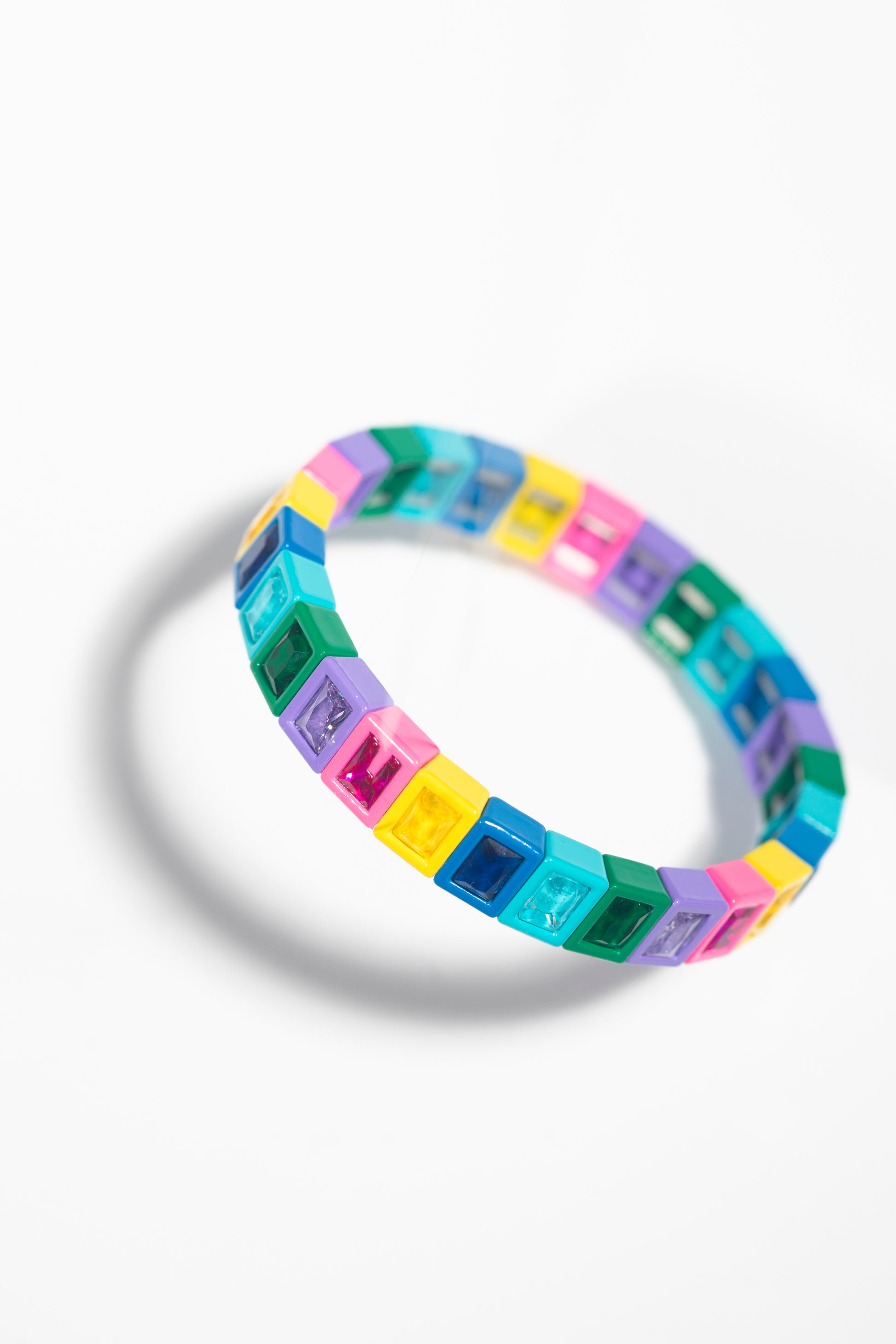 A colorful tile bracelet photographed at an angle against a white background with soft shadows. The bracelet is made of square plastic tiles with gem-like insets in rainbow colors including pink, yellow, blue, green, purple, and turquoise. The angled perspective emphasizes the three-dimensional nature of the tiles and their reflective surfaces.