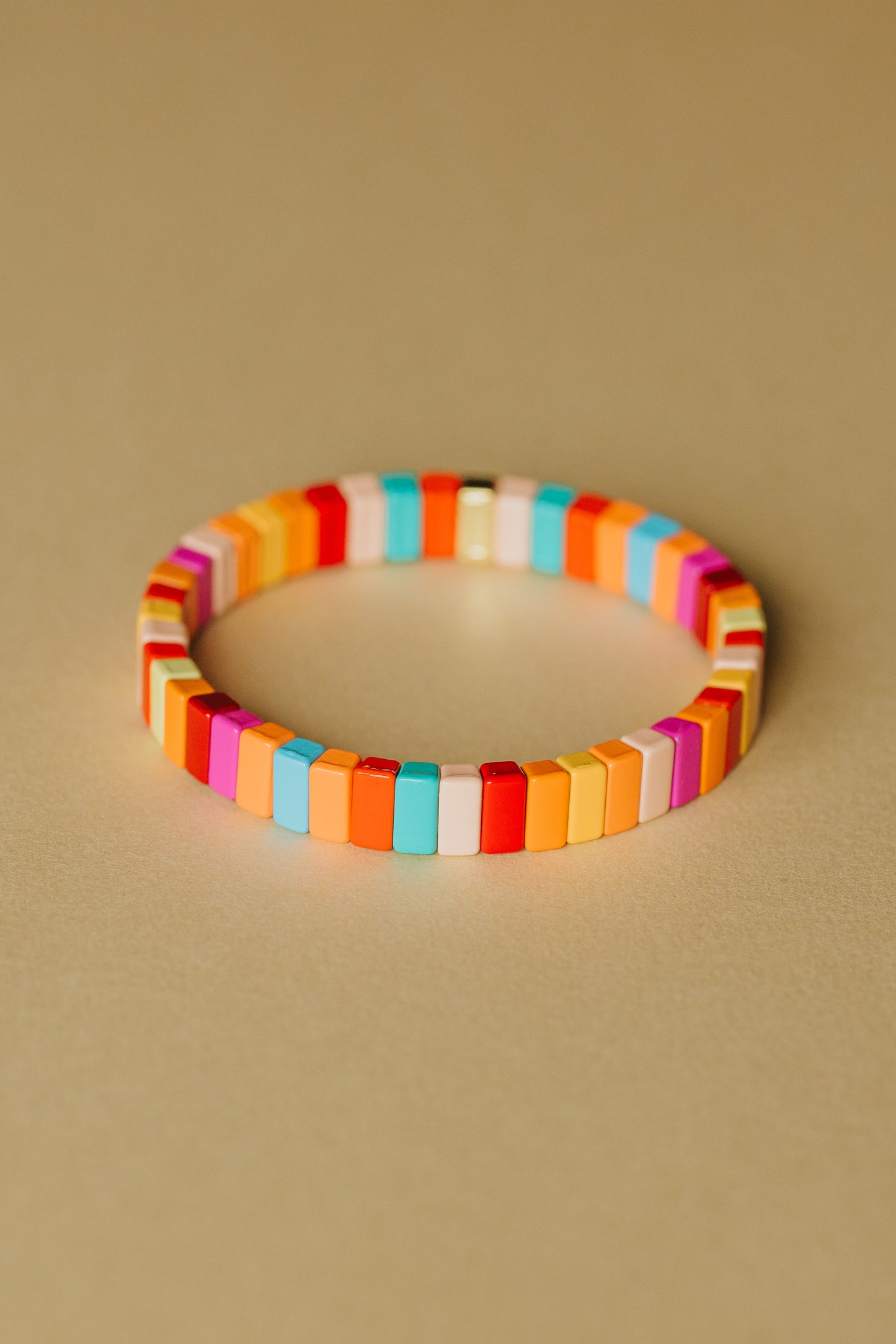 A single elastic tile bracelet photographed straight-on against a beige background. The bracelet is composed of uniform rectangular tiles arranged in a repeating pattern of bright summer colors including turquoise, coral, orange, yellow, pink, red, and white. The tiles create a continuous band with a modern, minimal aesthetic.