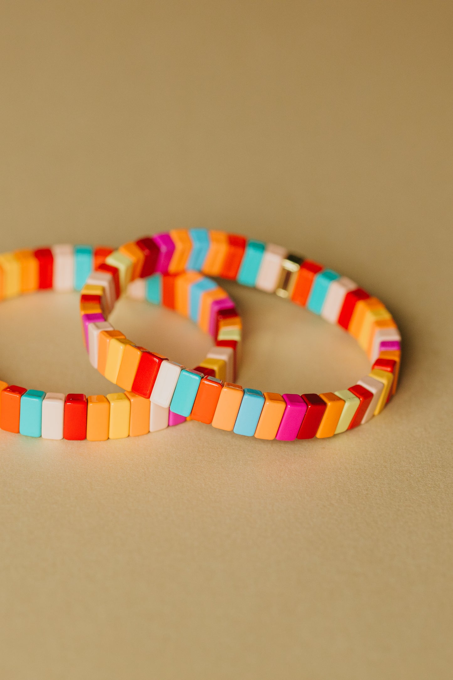 Two overlapping elastic tile bracelets photographed against a warm beige background. Each bracelet is made of rectangular tiles in a summer-inspired color palette including turquoise, coral, orange, yellow, pink, red, and white. The bracelets cast subtle shadows on the surface.