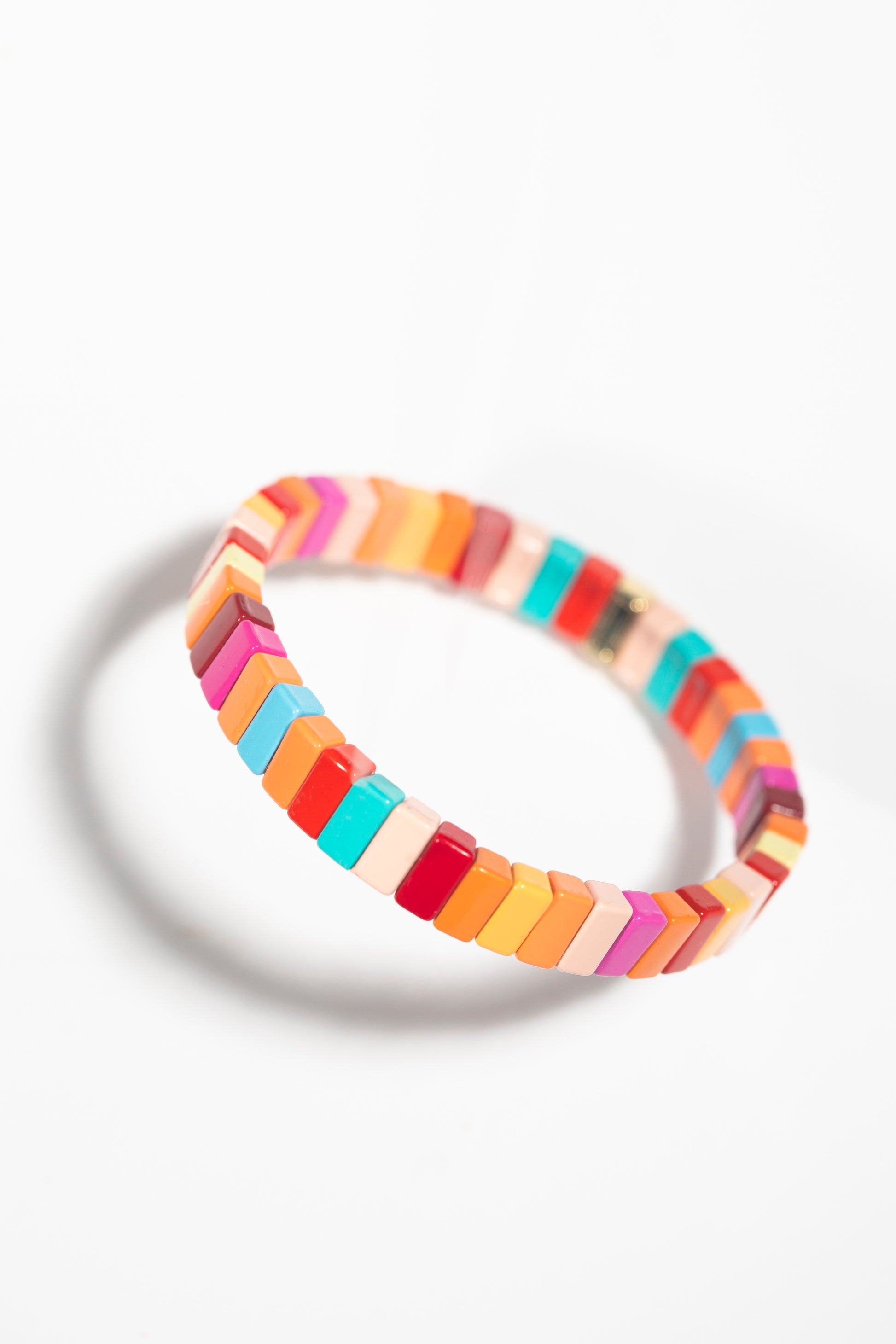 A tile bracelet photographed at an angled perspective against a white background, creating a dynamic shadow. The rectangular tiles are arranged in a gradient-like pattern of warm and cool tones including turquoise, coral, orange, yellow, pink, red, and white. The angled view emphasizes the three-dimensional quality of the individual tiles.