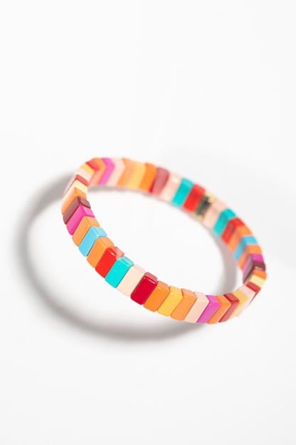 A tile bracelet photographed at an angled perspective against a white background, creating a dynamic shadow. The rectangular tiles are arranged in a gradient-like pattern of warm and cool tones including turquoise, coral, orange, yellow, pink, red, and white. The angled view emphasizes the three-dimensional quality of the individual tiles.