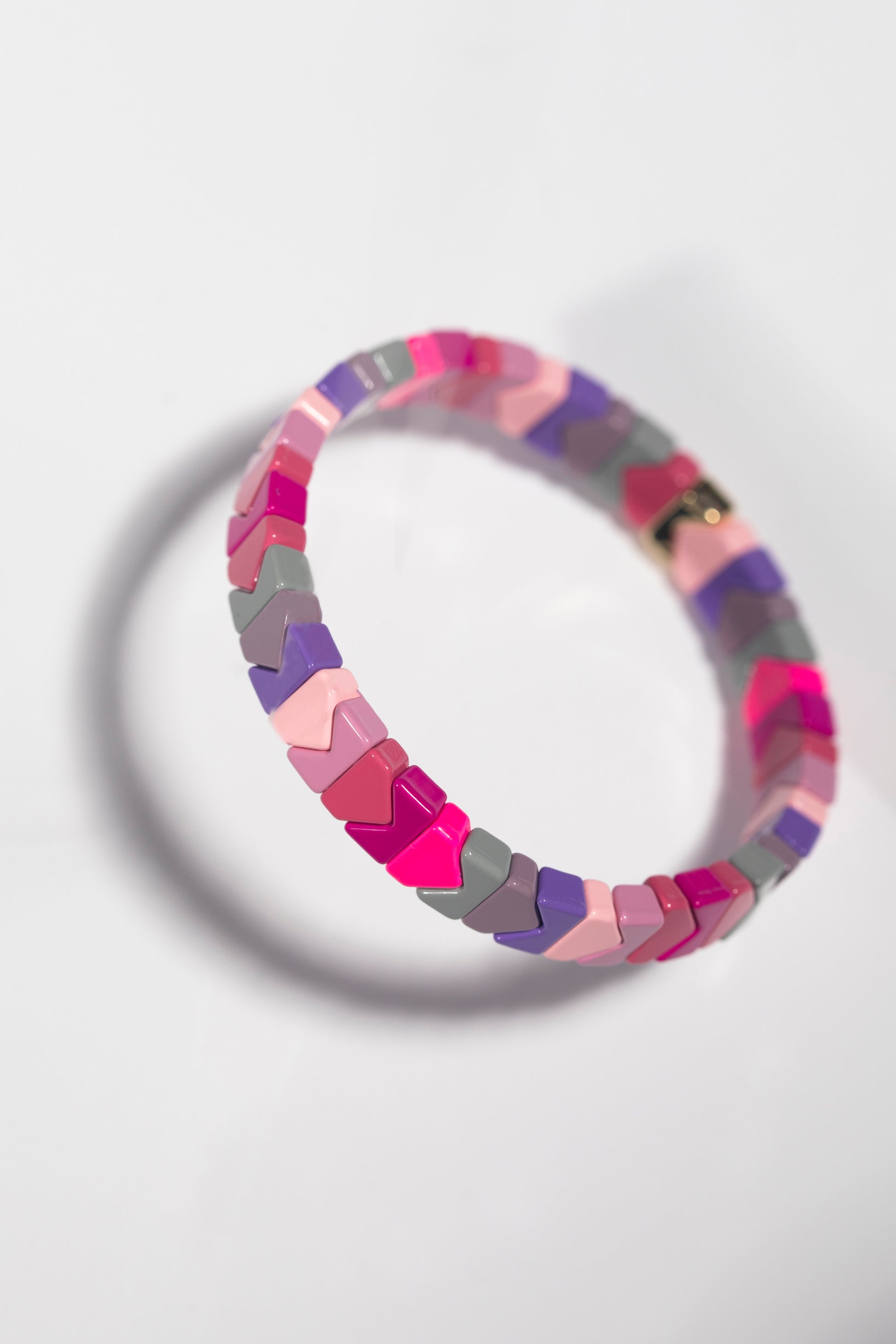 The same tile bracelet photographed at an angled perspective against a white background, casting a distinct shadow. The bracelet's tiles appear to be chevron or arrow-shaped from this angle, creating an interesting visual effect with the alternating colors of hot pink, light pink, purple, grey, and mauve. The angled view highlights the glossy finish of the tiles.