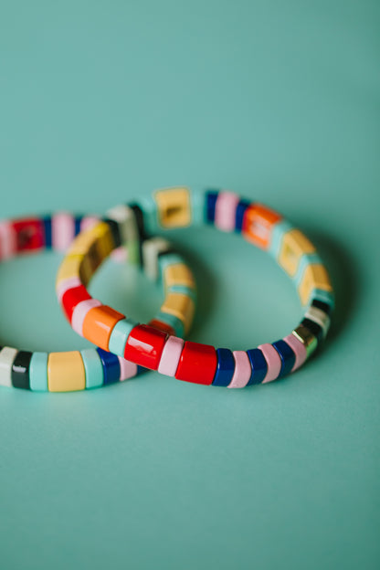 Two overlapping tile bracelets photographed against a mint green background. The bracelets feature a pattern of rectangular tiles in bright colors including yellow, red, navy blue, mint green, pink, orange, and black. The shallow depth of field creates an artistic blur effect.