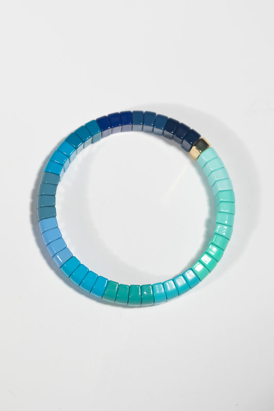 A tile bracelet photographed against a white background, featuring a beautiful gradient of blue and mint green tones. The rectangular tiles transition smoothly from navy blue to turquoise to mint green, creating an ombré effect around the circular bracelet. The tiles have a glossy, pearlescent finish that catches the light, and there appears to be a single gold or metallic tile connecting the color gradient. The bracelet casts a soft shadow, emphasizing its three-dimensional form.