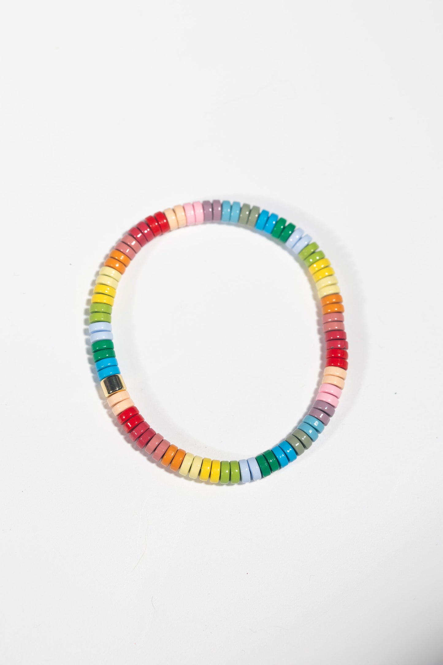 A circular elastic bracelet photographed straight-on against a white background. The bracelet features small disc-shaped tiles arranged in a rainbow gradient pattern that flows through the entire color spectrum. The colors transition smoothly from red to orange, yellow, green, blue, and back to red, with subtle pastel variations between each main color. A small gold connector piece joins the ends of the bracelet. 